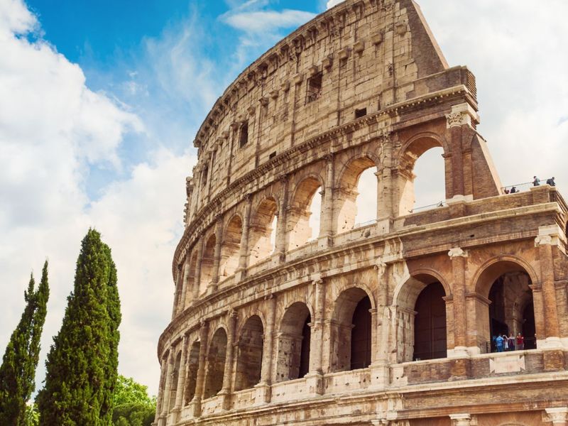 The Colosseum in Rome against a blue sky with clouds, flanked by green cypress trees.