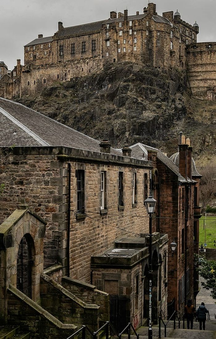 Historic stone buildings and a narrow street lead to a large castle atop a rocky hill under a cloudy sky.