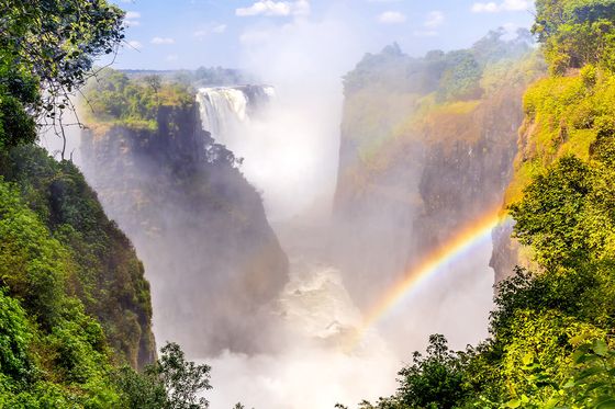 A majestic waterfall cascades into a verdant gorge, with mist creating a vivid rainbow under a bright, blue sky.