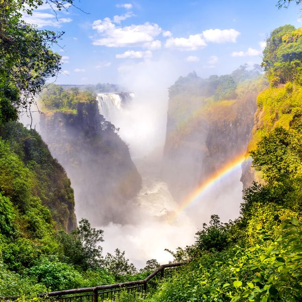 A majestic waterfall cascades into a verdant gorge, with mist creating a vivid rainbow under a bright, blue sky.