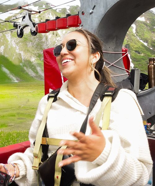 Woman in sunglasses and sweater smiles while preparing for a zipline ride in a mountainous area with green hills in the background.