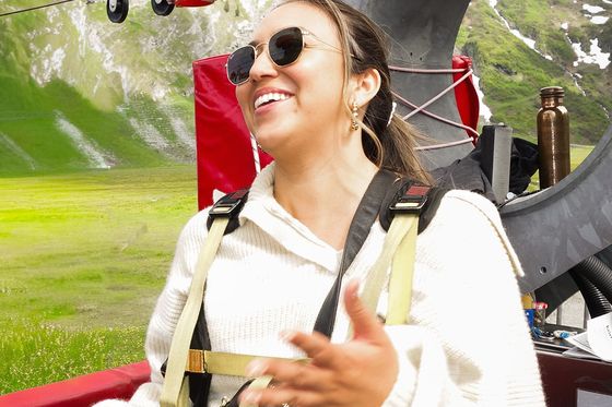 Woman in sunglasses and sweater smiles while preparing for a zipline ride in a mountainous area with green hills in the background.