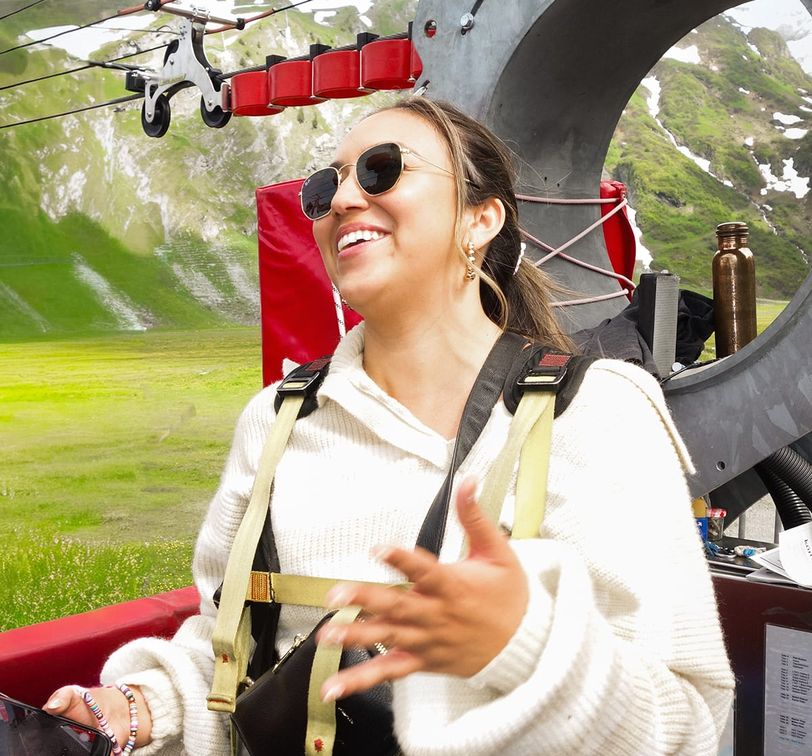A woman wearing sunglasses and a harness smiles while standing outdoors, with a grassy landscape and mountains in the background.