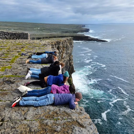 People lying on a rocky cliff edge overlooking the ocean, wearing casual clothes, with a rugged coastline and cloudy sky in the background.
