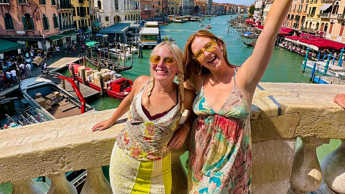Two women smiling and posing on a bridge over a canal in a sunny cityscape with colorful buildings and boats.