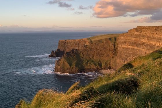 Cliffside landscape with green grass, rugged cliffs, and ocean waves under a partly cloudy sky at sunset.