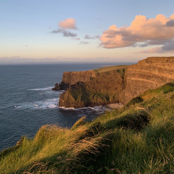 Cliffside landscape with green grass, rugged cliffs, and ocean waves under a partly cloudy sky at sunset.