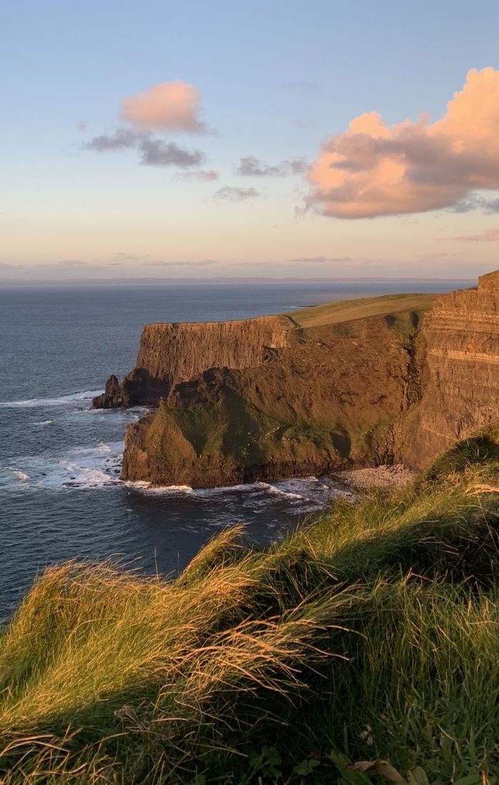 Cliffside landscape with green grass, rugged cliffs, and ocean waves under a partly cloudy sky at sunset.