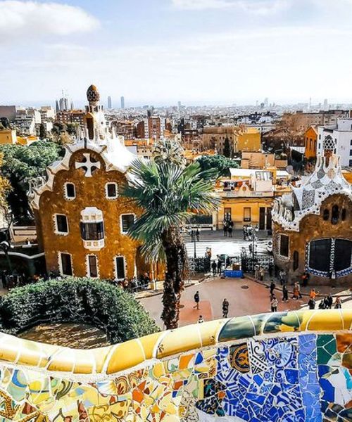 Colorful mosaic tiles and whimsical buildings in Park Güell, Barcelona, with a cityscape view in the background under a partly cloudy sky.