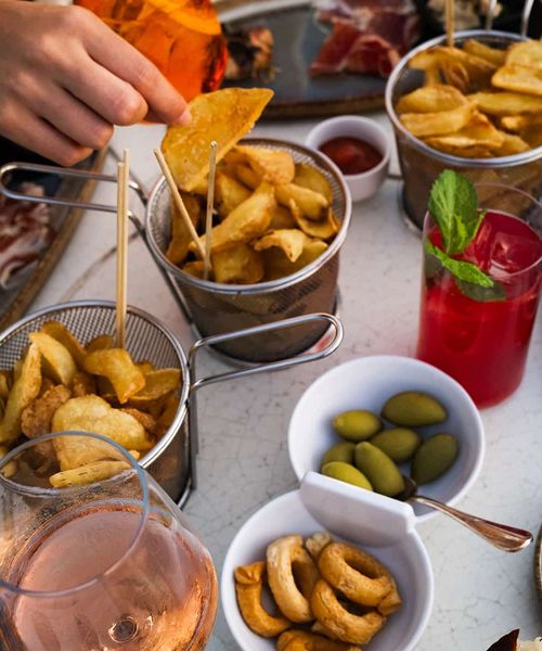 A variety of snacks including crisps, olives, and breadsticks on a table. A hand dips a crisp. Red drink with mint and a glass of rosé nearby.