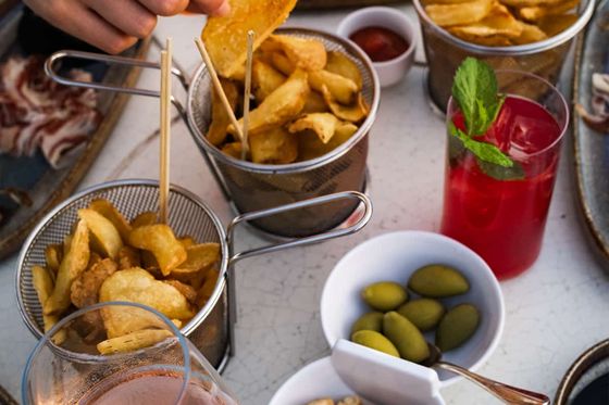 A variety of snacks including crisps, olives, and breadsticks on a table. A hand dips a crisp. Red drink with mint and a glass of rosé nearby.