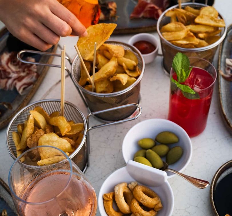 A variety of snacks including crisps, olives, and breadsticks on a table. A hand dips a crisp. Red drink with mint and a glass of rosé nearby.