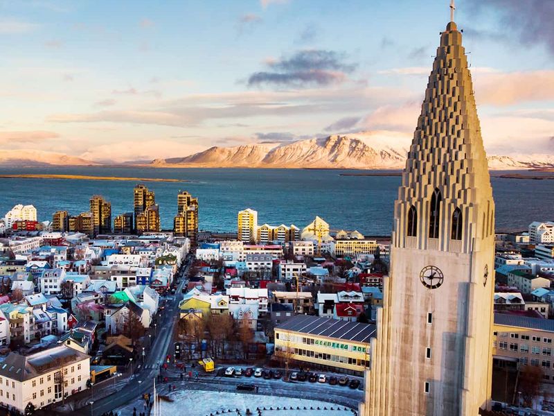 Aerial view of Reykjavik with Hallgrímskirkja church tower, colorful buildings, and distant snow-capped mountains under a partly cloudy sky.