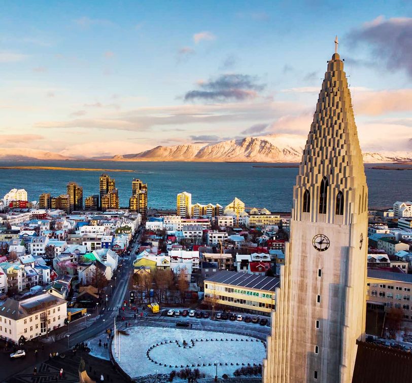 Aerial view of a cityscape with a tall, pointed church tower, colorful buildings, distant mountains, and a body of water during sunset.