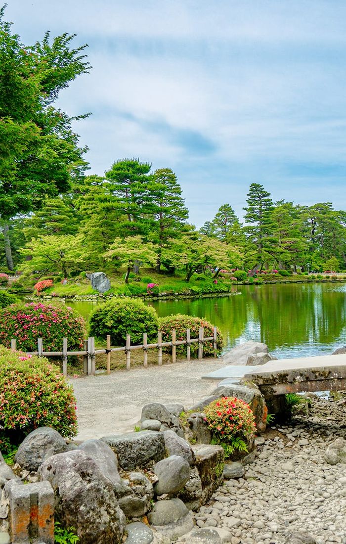 A serene Japanese garden with a pond, stone bridge, lush greenery, and colorful shrubs under a clear blue sky.