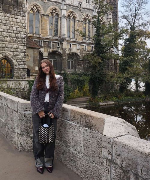Woman in a gray sweater and jeans stands on a stone bridge beside a historic building and river, with trees in the background.