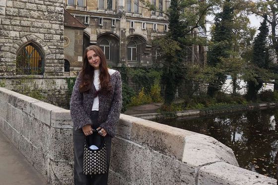 Woman in a gray sweater and jeans stands on a stone bridge beside a historic building and river, with trees in the background.
