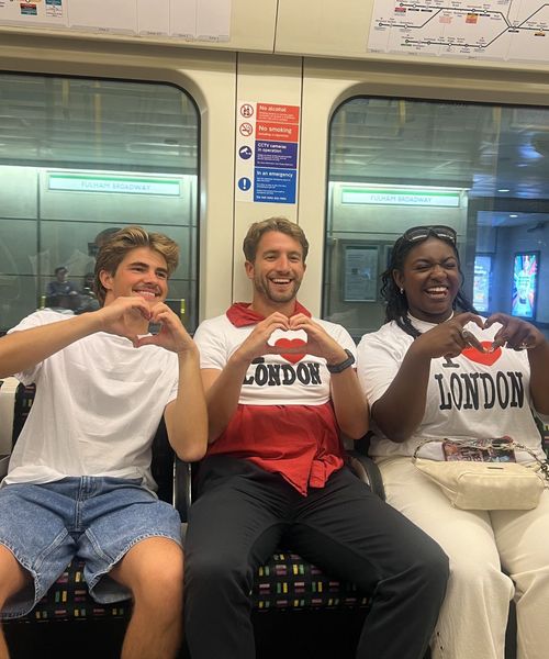 Three people sitting on a subway wearing "I Love London" shirts, making heart shapes with their hands, and smiling.
