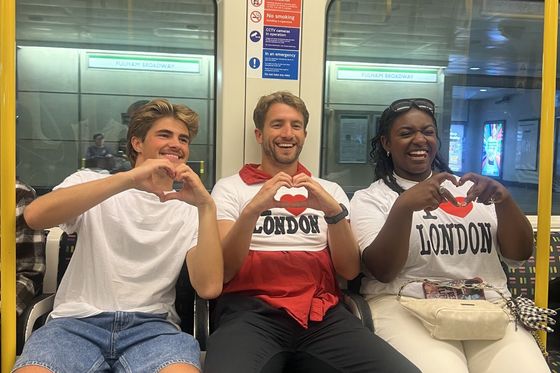 Three people sitting on a subway wearing "I Love London" shirts, making heart shapes with their hands, and smiling.