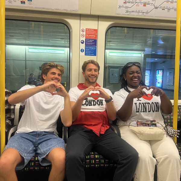Three people sitting on a subway wearing "I Love London" shirts, making heart shapes with their hands, and smiling.