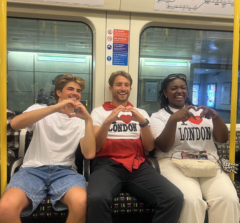 Three people sitting on a subway wearing "I Love London" shirts, making heart shapes with their hands, and smiling.