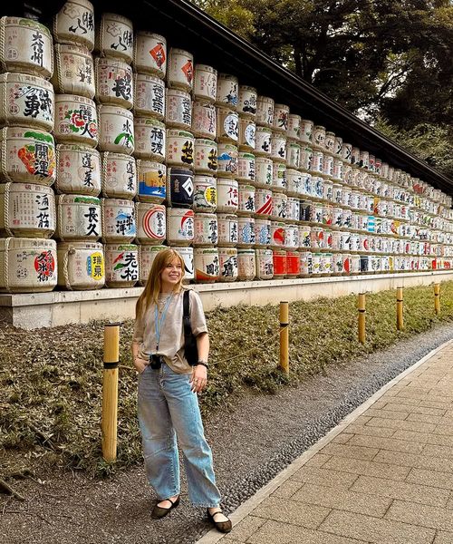 A person stands smiling in front of a wall of stacked sake barrels with colorful designs, surrounded by trees and a paved path.