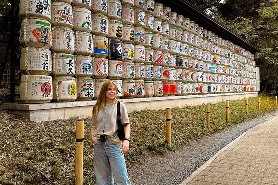 A person stands smiling in front of a wall of stacked sake barrels with colorful designs, surrounded by trees and a paved path.