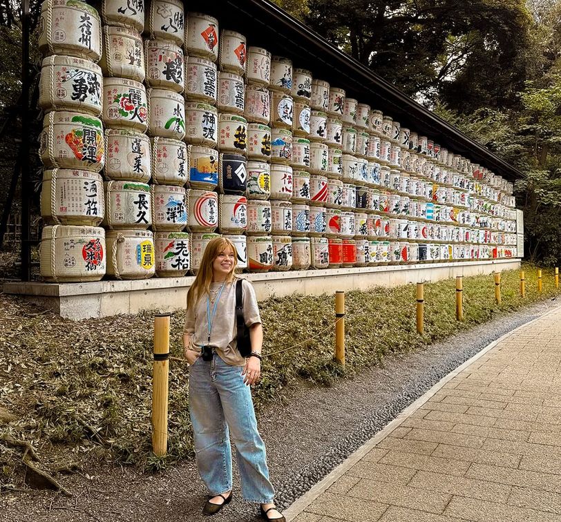 A person stands smiling in front of a wall of stacked sake barrels with colorful designs, surrounded by trees and a paved path.