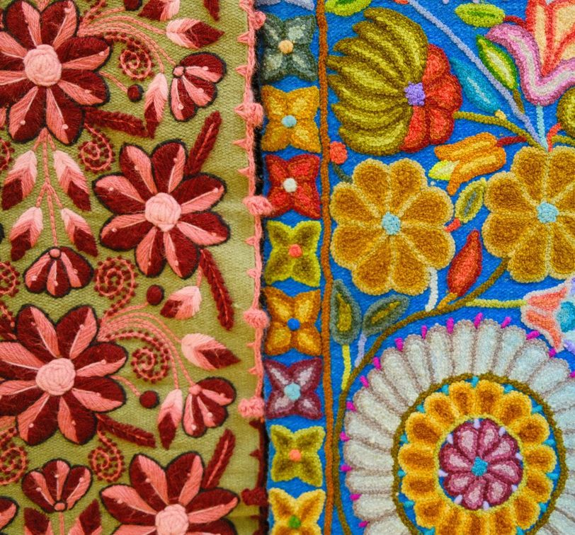 Colorful embroidery featuring intricate floral patterns with pink, red, and yellow flowers on green and blue fabric backgrounds.