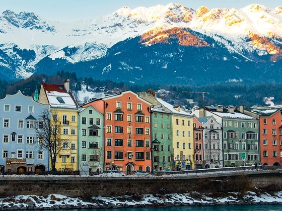 Colorful buildings line a riverbank, with snow-covered mountains in the background, under a clear blue sky.