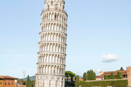The Leaning Tower of Pisa, a white marble bell tower, leans prominently in a grassy area with tourists nearby under a clear blue sky.