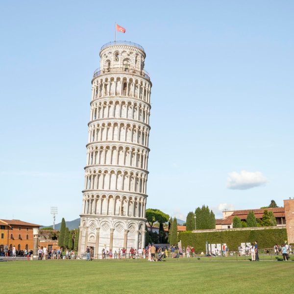 The Leaning Tower of Pisa, a white marble bell tower, leans prominently in a grassy area with tourists nearby under a clear blue sky.