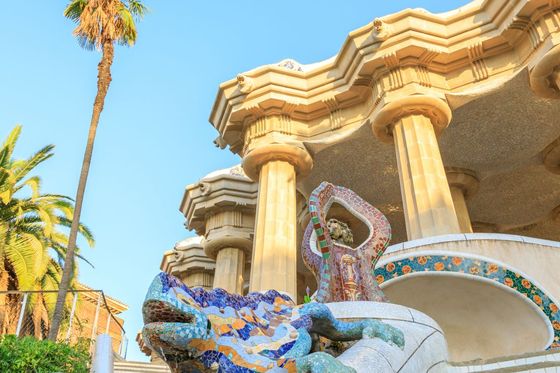 Colorful mosaic lizard sculpture on stairs in Park Güell, Barcelona, with columns and palm trees in the background under a clear blue sky.