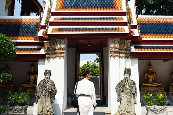 A person in white attire walks towards a colorful traditional temple entrance flanked by statues, golden Buddhas, and greenery.