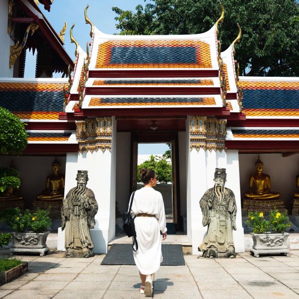 A person in white attire walks towards a colorful traditional temple entrance flanked by statues, golden Buddhas, and greenery.