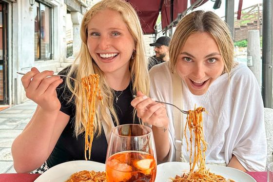 Two women smile while enjoying spaghetti at an outdoor restaurant. A glass with an orange drink sits on the table in front of them.