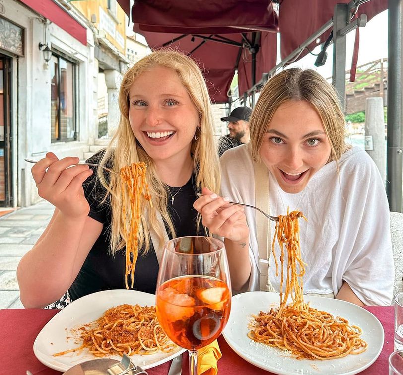 Two women smile while enjoying spaghetti at an outdoor restaurant. A glass with an orange drink sits on the table in front of them.