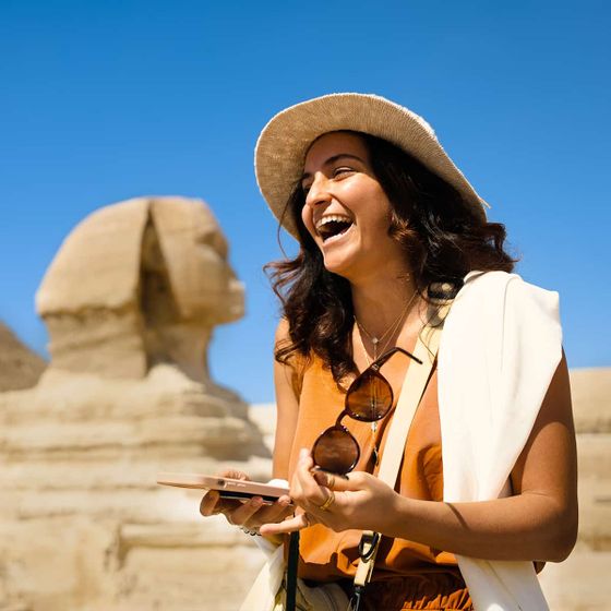 A woman in a sunhat laughs joyfully in front of the Sphinx under a clear blue sky, holding a smartphone and sunglasses.