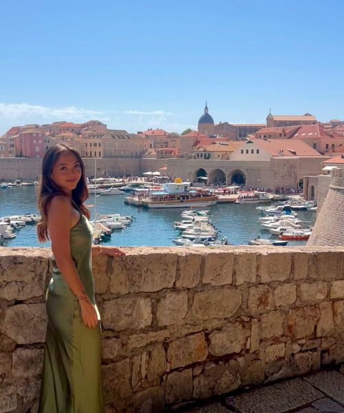 Woman in a green dress stands by a stone wall, overlooking a marina with boats and historic buildings in the background under a clear blue sky.