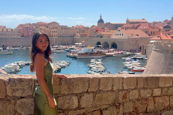 Woman in a green dress stands by a stone wall, overlooking a marina with boats and historic buildings in the background under a clear blue sky.