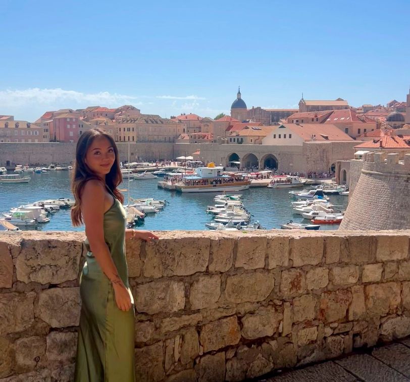 Woman in a green dress stands by a stone wall, overlooking a marina with boats and historic buildings in the background under a clear blue sky.