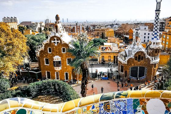 View of Park Güell in Barcelona with colorful mosaics, whimsical buildings, and a distant cityscape under a partly cloudy sky.