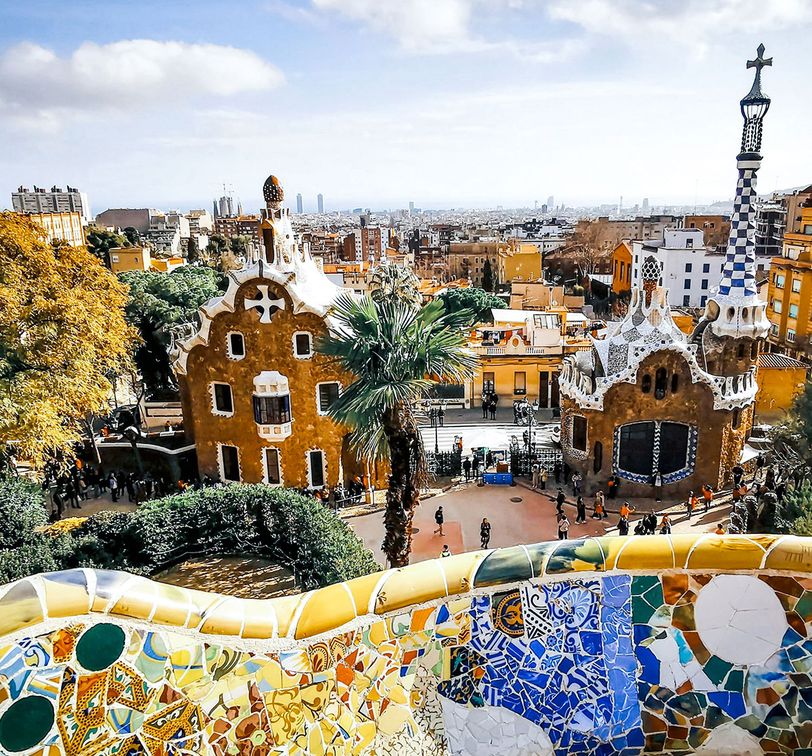 View of Park Güell in Barcelona with colorful mosaics, whimsical buildings, and a distant cityscape under a partly cloudy sky.