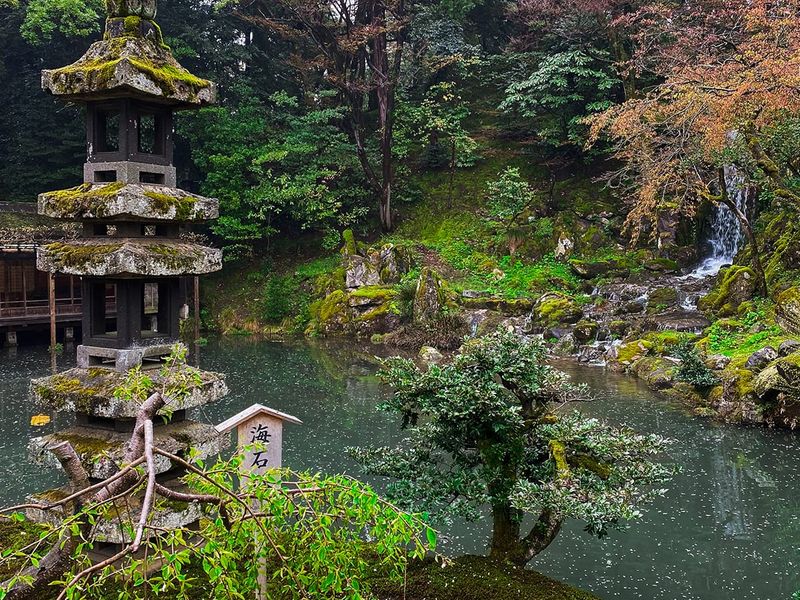 A serene Japanese garden with a stone lantern, lush greenery, a small waterfall, and a tranquil pond surrounded by trees.