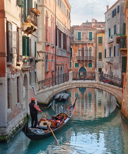 Gondolier navigating a canal in Venice, Italy, with historical buildings and a stone bridge in the background on a sunny day.