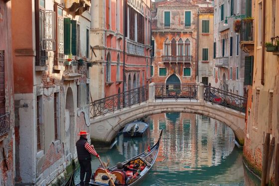 Gondolier navigating a canal in Venice, Italy, with historical buildings and a stone bridge in the background on a sunny day.
