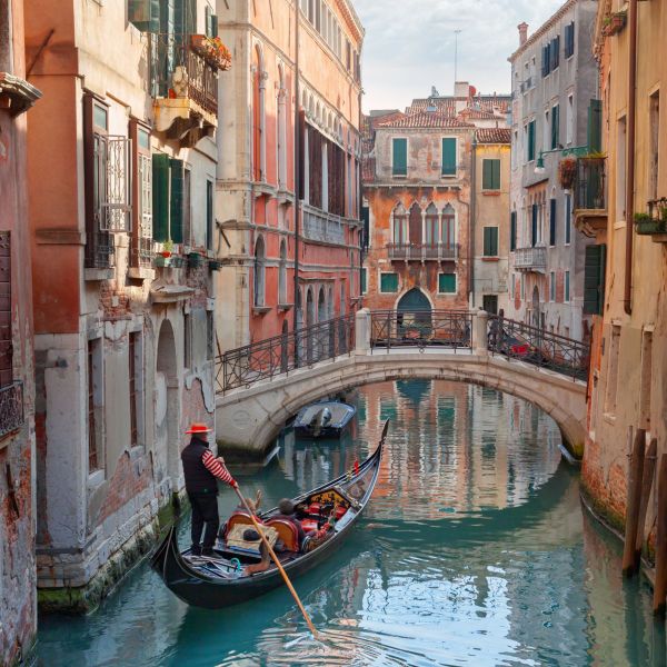 Gondolier navigating a canal in Venice, Italy, with historical buildings and a stone bridge in the background on a sunny day.