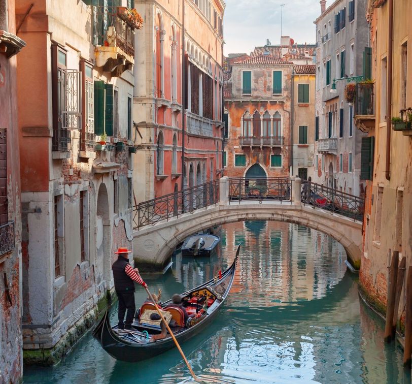 Gondolier navigating a canal in Venice, Italy, with historical buildings and a stone bridge in the background on a sunny day.