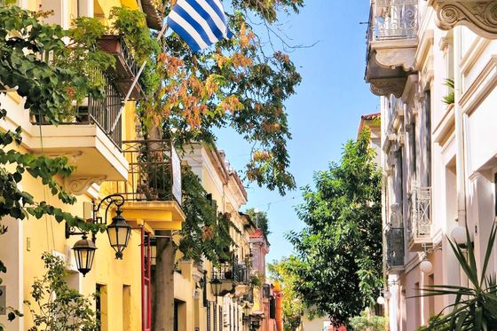 A picturesque, narrow street in Greece, lined with yellow and white buildings, lush trees, and a Greek flag waving from a balcony.