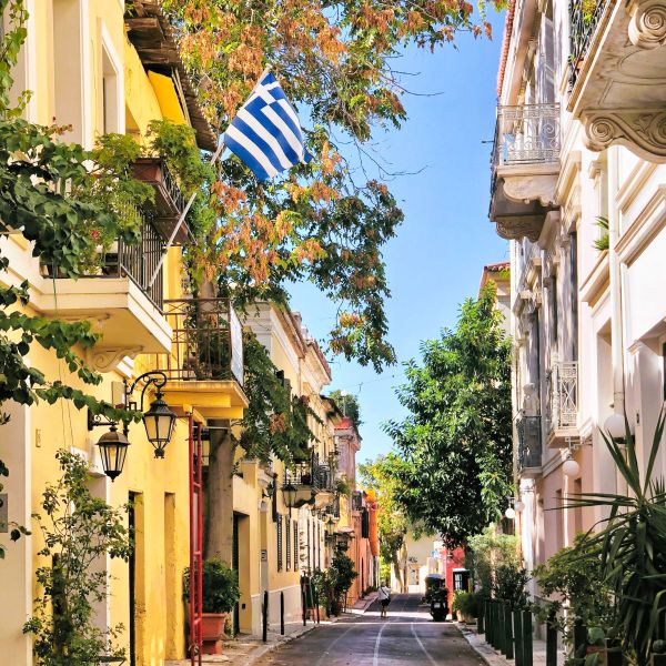 A picturesque, narrow street in Greece, lined with yellow and white buildings, lush trees, and a Greek flag waving from a balcony.